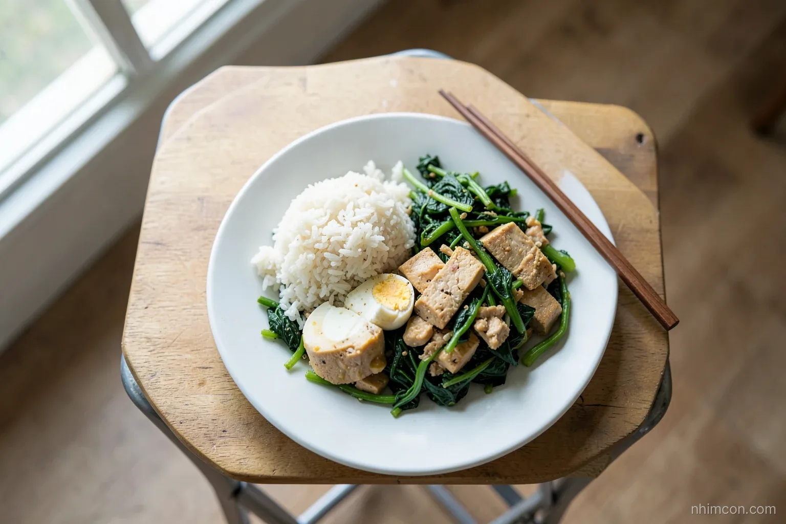 Overhead shot of a simple plate on a small folding table: rice, stir-fried greens, tofu and a boiled egg, chopsticks resting, natural window light