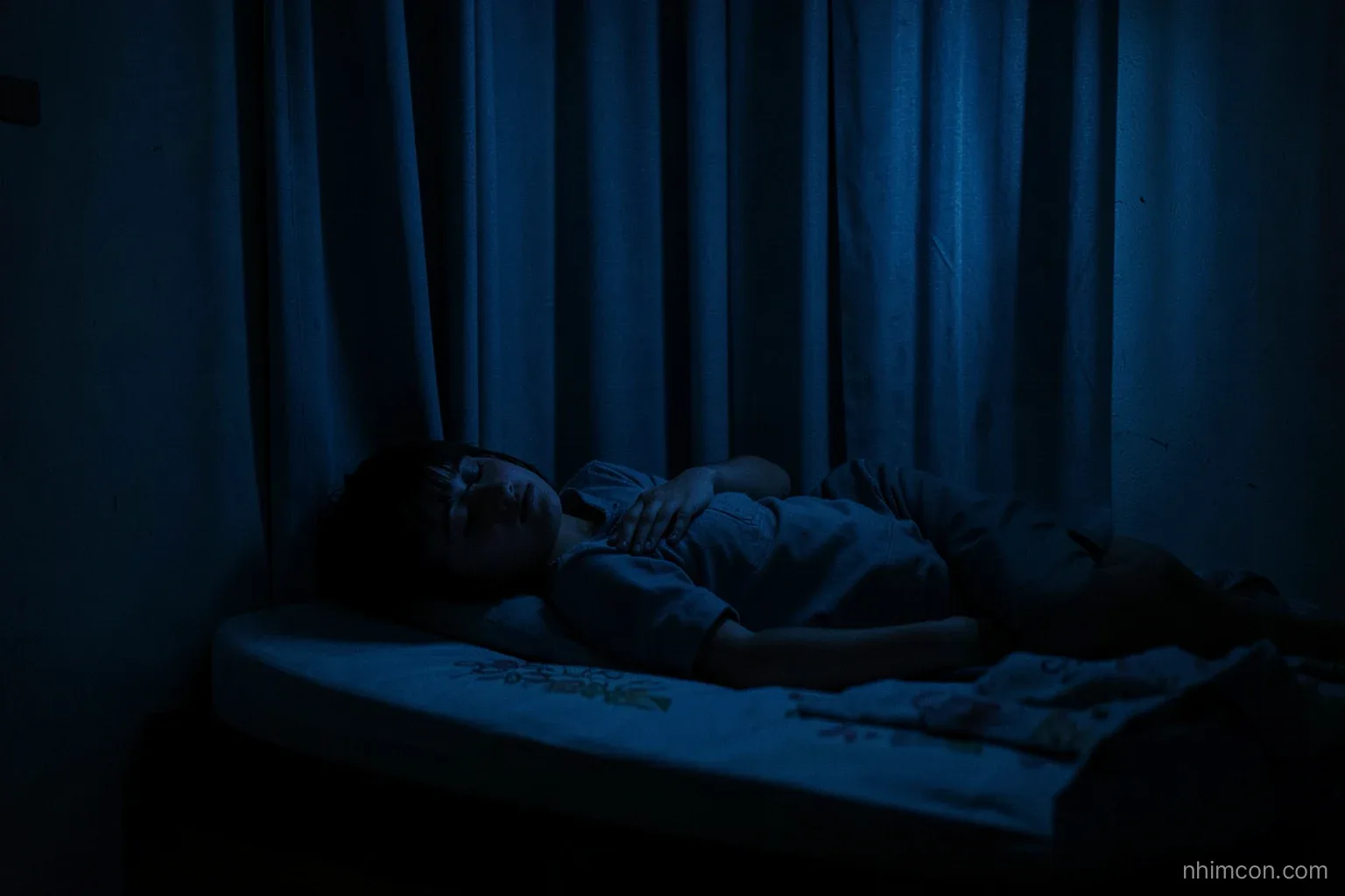 Dim small room at night, a young person asleep facing the wall on a low mattress, thick curtain drawn, quiet blue tones